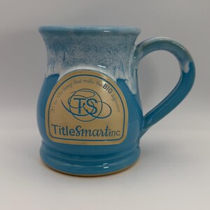May include: A blue ceramic mug with a white drip glaze at the rim. The front features a tan label with the text "It's the little things that make the BIG difference", a "TS" logo, and "TitleSmartinc" printed in blue ink on the side of the mug.