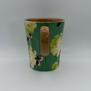 Puede incluir: Una taza de cer&aacute;mica con fondo verde y un estampado floral en tonos blancos y verdes. La taza tiene interior marr&oacute;n y asa marr&oacute;n. El dise&ntilde;o floral presenta rosas y otras flores.