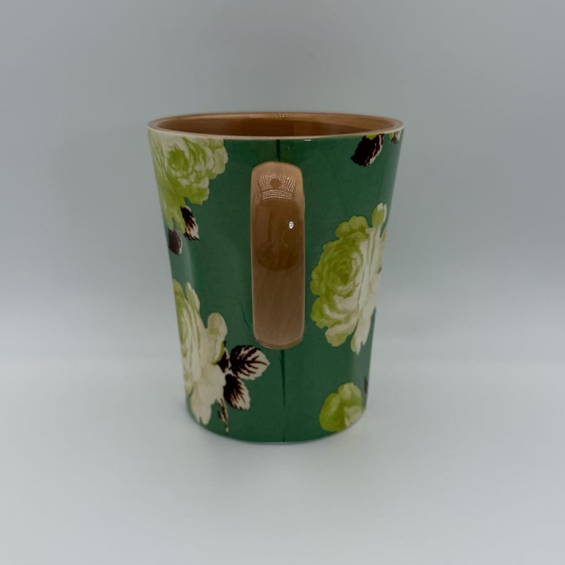 Puede incluir: Una taza de cer&aacute;mica con fondo verde y un estampado floral en tonos blancos y verdes. La taza tiene interior marr&oacute;n y asa marr&oacute;n. El dise&ntilde;o floral presenta rosas y otras flores.