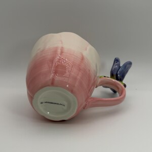 May include: A ceramic mug with a pink and white gradient design. The handle is pink and features a blue floral accent. The bottom of the mug has the text "by ANTHROPOLOGIE".