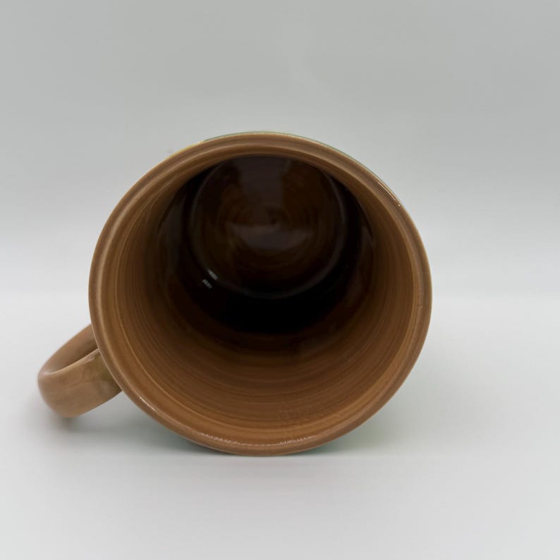 Puede incluir: Una taza de cer&aacute;mica marr&oacute;n con asa, vista desde arriba. El interior es de un tono marr&oacute;n m&aacute;s oscuro, y el exterior tiene un aspecto texturizado y terroso. La taza est&aacute; sobre un fondo blanco liso.