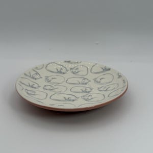 May include: A small, round ceramic plate with a cream-colored surface. The plate is decorated with a repeating pattern of sleeping cat illustrations in a muted blue-gray color. The rim of the plate is a reddish-brown color.