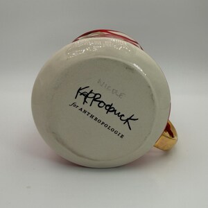 Pode incluir: Uma caneca de cer&acirc;mica branca com uma al&ccedil;a dourada e detalhes vermelhos. A parte inferior da caneca apresenta o texto "KP Rodruck for ANTHROPOLOGIE" em escrita preta. A caneca tem formato redondo e acabamento brilhante.