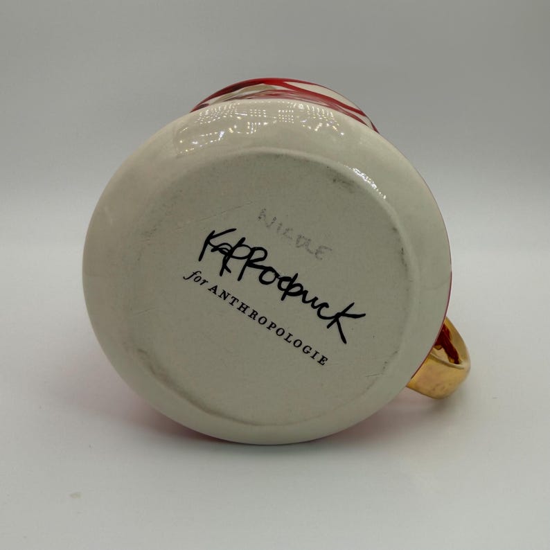 Pode incluir: Uma caneca de cer&acirc;mica branca com uma al&ccedil;a dourada e detalhes vermelhos. A parte inferior da caneca apresenta o texto "KP Rodruck for ANTHROPOLOGIE" em escrita preta. A caneca tem formato redondo e acabamento brilhante.