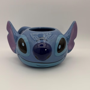 Puede incluir: Una taza de cer&aacute;mica con la forma del personaje Stitch de la pel&iacute;cula de Disney Lilo & Stitch. La taza es de color azul claro con ojos negros grandes, una nariz azul oscuro y orejas moradas. Tiene un asa y una abertura ancha.