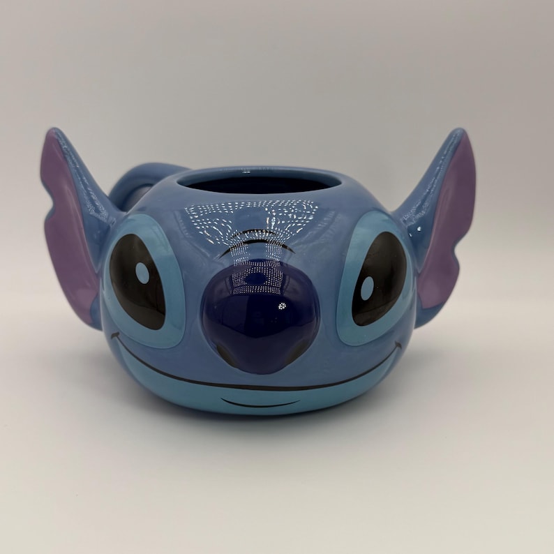 Puede incluir: Una taza de cer&aacute;mica con la forma del personaje Stitch de la pel&iacute;cula de Disney Lilo & Stitch. La taza es de color azul claro con ojos negros grandes, una nariz azul oscuro y orejas moradas. Tiene un asa y una abertura ancha.