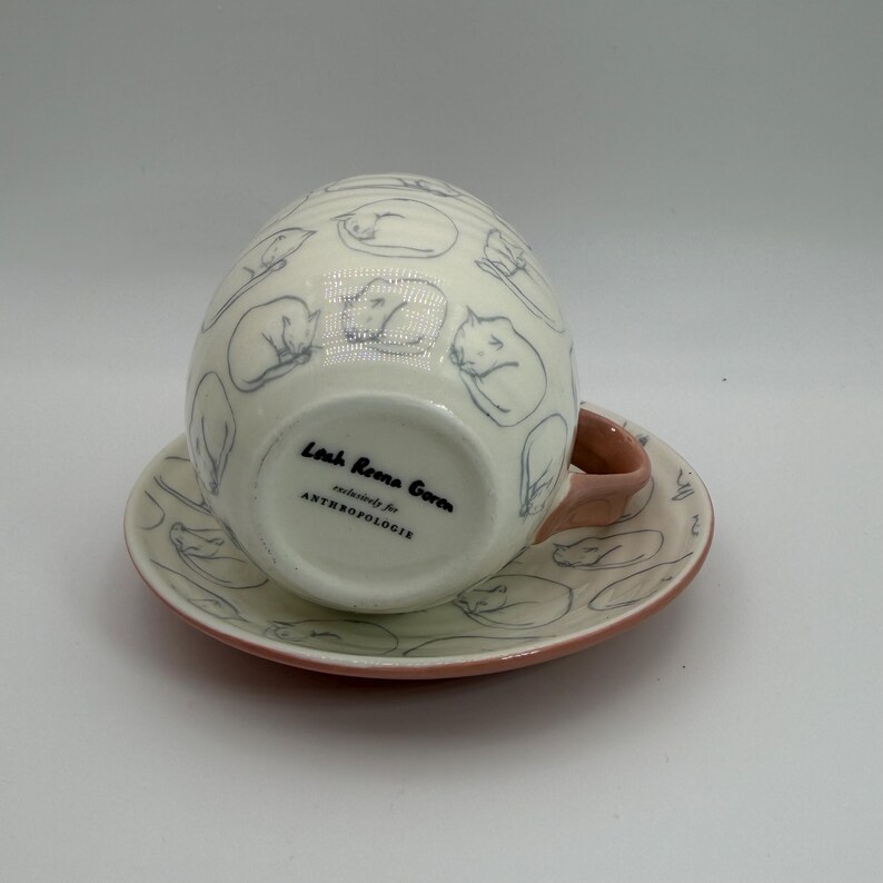 May include: A white ceramic teacup and saucer set featuring a repeating pattern of sleeping cats in blue. The cup has a pink handle, and the saucer has a pink rim. The bottom of the cup reads "Leah Reena Goren exclusively for ANTHROPOLOGIE."