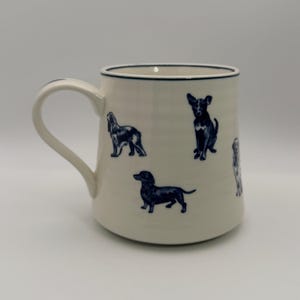 K&ouml;nnte beinhalten: Cremefarbene Keramiktasse mit marineblauen Illustrationen verschiedener Hunderassen. Die Tasse hat einen gebogenen Henkel und einen marineblauen Rand. Die Hunde sind in verschiedenen Posen dargestellt, was dem Design eine spielerische Note verleiht.