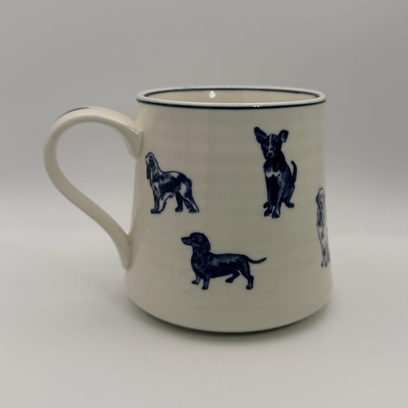 K&ouml;nnte beinhalten: Cremefarbene Keramiktasse mit marineblauen Illustrationen verschiedener Hunderassen. Die Tasse hat einen gebogenen Henkel und einen marineblauen Rand. Die Hunde sind in verschiedenen Posen dargestellt, was dem Design eine spielerische Note verleiht.