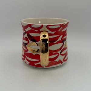Pode incluir: Uma caneca de cer&acirc;mica com fundo branco, coberta por um padr&atilde;o de l&aacute;bios vermelhos. A caneca tem uma al&ccedil;a dourada e detalhes dourados nos desenhos dos l&aacute;bios. O interior da caneca &eacute; creme.