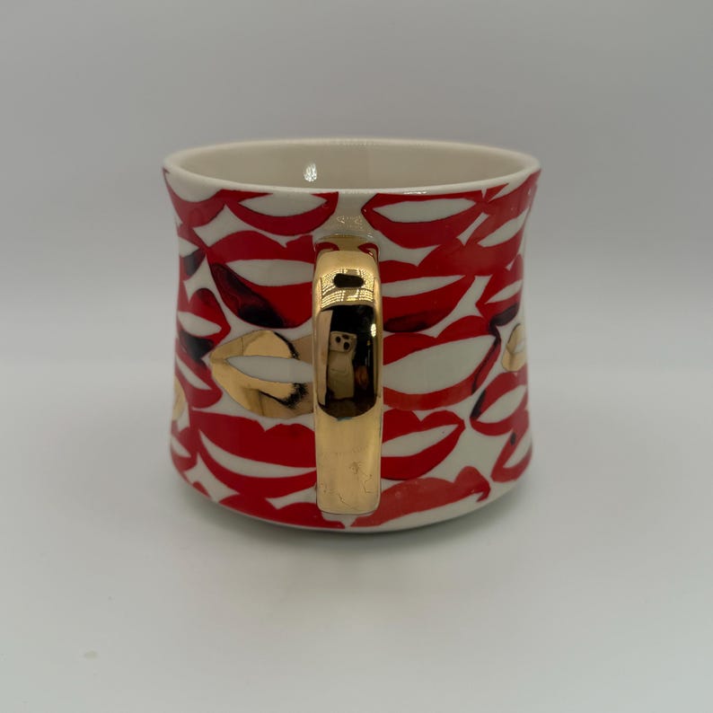 Pode incluir: Uma caneca de cer&acirc;mica com fundo branco, coberta por um padr&atilde;o de l&aacute;bios vermelhos. A caneca tem uma al&ccedil;a dourada e detalhes dourados nos desenhos dos l&aacute;bios. O interior da caneca &eacute; creme.