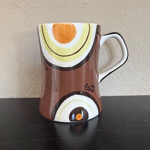 Könnte beinhalten: Eine Keramik-Kaffeetasse mit einem braunen, weißen, gelben und orangefarbenen Design. Die Tasse hat einen weißen Griff mit schwarzem Rand. Das Wort "Latte" steht auf der Seite der Tasse.