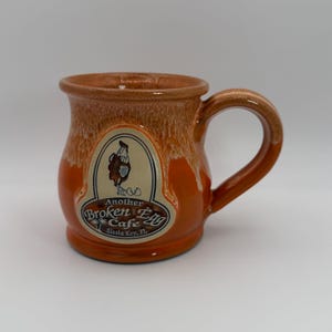 Op de afbeelding: Een keramische mok met een afgeronde body en een grote handgreep. De mok is voornamelijk oranje met een bruine rand en handgreep. Een crèmekleurig ovaal etiket toont een illustratie van een haan en de tekst "Another Broken Egg Cafe, Siesta Key, FL."