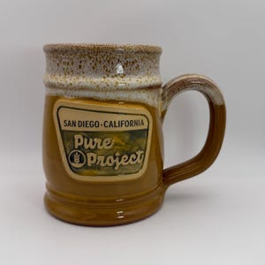 May include: A ceramic mug with a glossy mustard yellow base and a speckled white rim. A central embossed label features the text SAN DIEGO CALIFORNIA and Pure Project with a wheat icon. The mug has a curved handle and a smooth, glazed finish.
