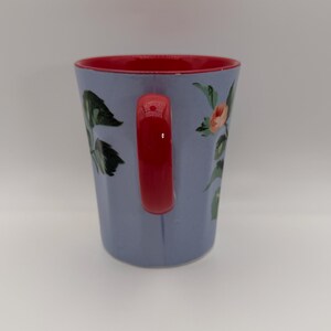 May include: A ceramic mug with a light blue exterior and a red interior and handle. The mug is decorated with floral designs in shades of pink, green, and white. The mug is designed for hot or cold beverages.