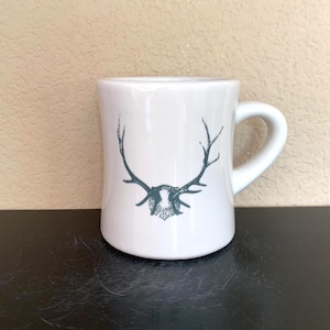 Puede incluir: Taza de café de cerámica blanca con asa curva. La taza presenta una ilustración verde oscuro de cornamentas de ciervo. La taza está sobre una superficie negra.
