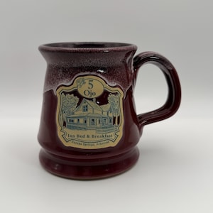 Puede incluir: Una taza de cerámica burdeos con asa y borde oscuros. La taza presenta una etiqueta color crema con el texto "5 Ojo Inn Bed & Breakfast Eureka Springs, Arkansas" y una ilustración de una casa.