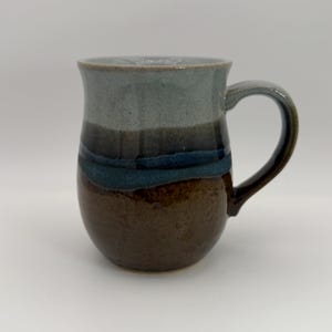 May include: A ceramic mug featuring a rounded body and a curved handle. The exterior displays a layered glaze design with a light gray top section, a dark blue middle band, and a textured brown base, creating a distinct horizontal color transition.