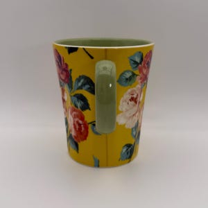 K&ouml;nnte beinhalten: Gelbe Keramiktasse mit einem Blumenmuster aus rosa, roten und wei&szlig;en Rosen und gr&uuml;nen Bl&auml;ttern. Der Innenraum ist gr&uuml;n und der Griff olivgr&uuml;n. Die Tasse hat eine Standardgr&ouml;&szlig;e f&uuml;r Hei&szlig;getr&auml;nke.