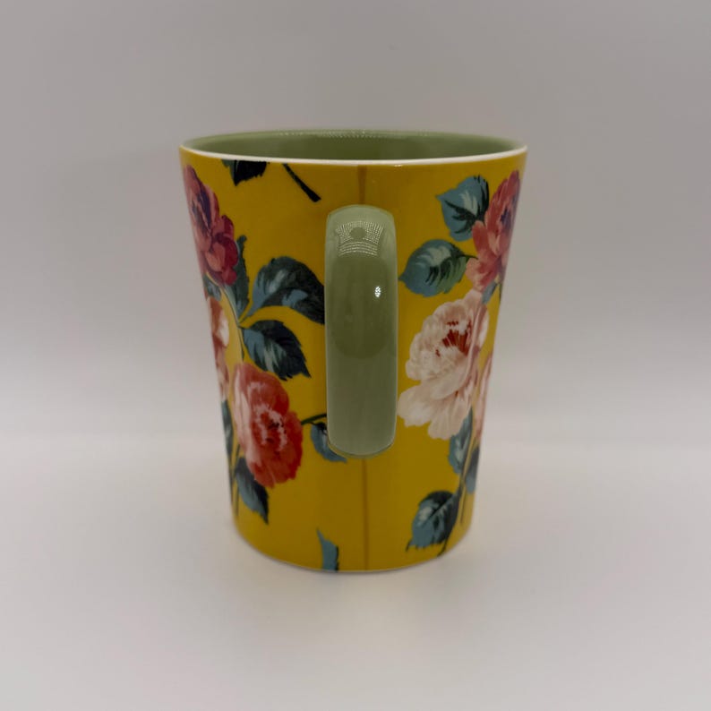 K&ouml;nnte beinhalten: Gelbe Keramiktasse mit einem Blumenmuster aus rosa, roten und wei&szlig;en Rosen und gr&uuml;nen Bl&auml;ttern. Der Innenraum ist gr&uuml;n und der Griff olivgr&uuml;n. Die Tasse hat eine Standardgr&ouml;&szlig;e f&uuml;r Hei&szlig;getr&auml;nke.