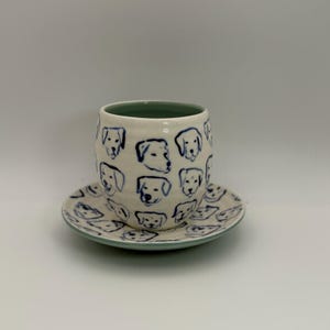 May include: A ceramic cup and saucer set. The cup and saucer are white with a blue dog face pattern. The rim of the cup is green. The saucer is round with a matching dog face pattern and a green rim.