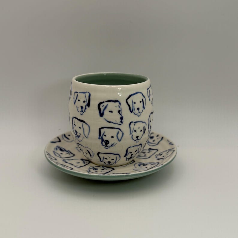 May include: A ceramic cup and saucer set. The cup and saucer are white with a blue dog face pattern. The rim of the cup is green. The saucer is round with a matching dog face pattern and a green rim.