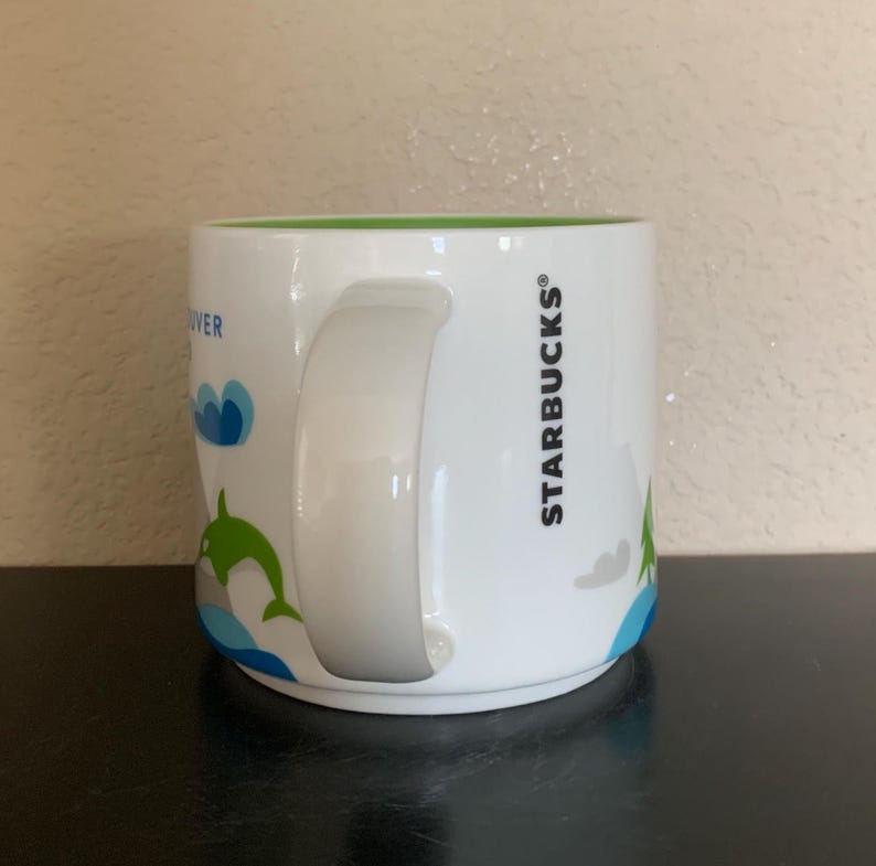 May include: White ceramic Starbucks mug with a green interior and a white handle. The mug features a blue and green graphic of a dolphin jumping out of the water, with the word "Vancouver" and the Starbucks logo.