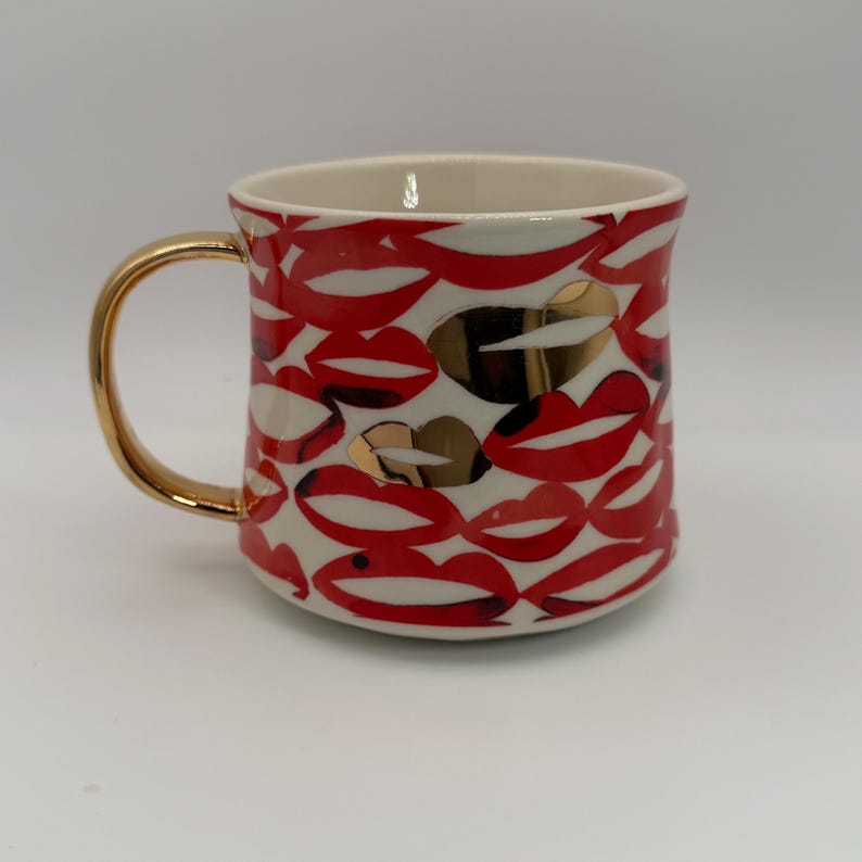 Pode incluir: Uma caneca de cer&acirc;mica com uma al&ccedil;a dourada e um padr&atilde;o de l&aacute;bios vermelhos sobre um fundo branco. Alguns l&aacute;bios s&atilde;o dourados. O interior da caneca &eacute; creme.