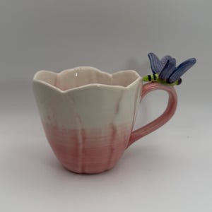 May include: A ceramic mug with a pink and white floral design. The mug has a handle and a decorative dragonfly on the rim. The mug is a light pink color at the bottom and fades to white at the top.