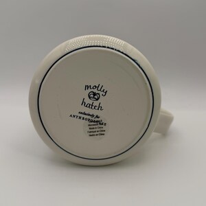K&ouml;nnte beinhalten: Eine wei&szlig;e Keramiktasse mit Henkel, mit dunkelblauem Rand und dem Text "molly hatch" mit einem Kronen-Design. Die Tasse ist exklusiv f&uuml;r Anthropologie und mikrowellengeeignet.