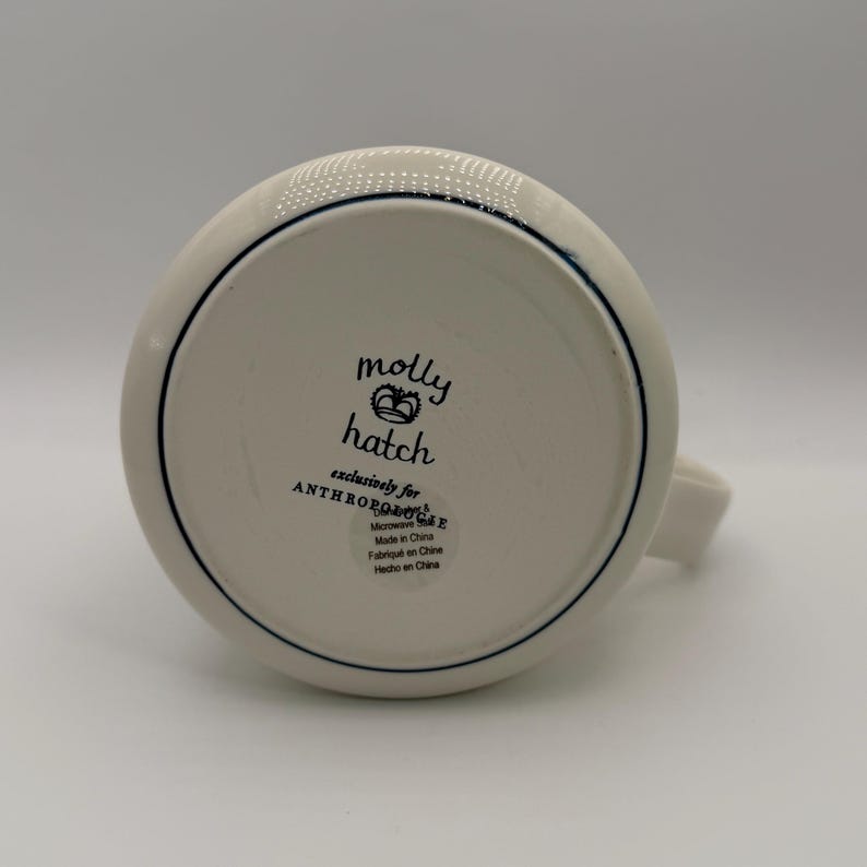 K&ouml;nnte beinhalten: Eine wei&szlig;e Keramiktasse mit Henkel, mit dunkelblauem Rand und dem Text "molly hatch" mit einem Kronen-Design. Die Tasse ist exklusiv f&uuml;r Anthropologie und mikrowellengeeignet.