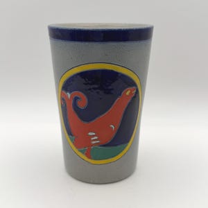 May include: A cylindrical gray ceramic tumbler featuring a dark blue rim and a central circular emblem. The emblem displays a stylized orange seal-like creature on a dark blue background, outlined in yellow, with a green base element.