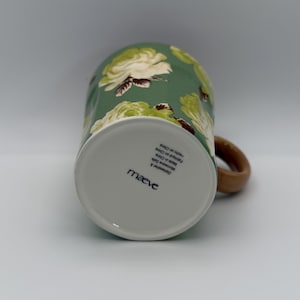Puede incluir: Una taza de cer&aacute;mica con fondo verde y un estampado floral de rosas blancas y verde claro. La taza tiene un asa marr&oacute;n y la parte inferior de la taza tiene el nombre de la marca "maeve" impreso. La taza es apta para lavavajillas y microondas.