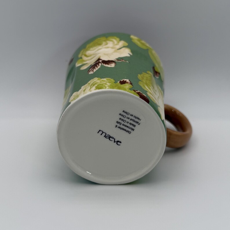 Puede incluir: Una taza de cer&aacute;mica con fondo verde y un estampado floral de rosas blancas y verde claro. La taza tiene un asa marr&oacute;n y la parte inferior de la taza tiene el nombre de la marca "maeve" impreso. La taza es apta para lavavajillas y microondas.