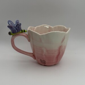 May include: A ceramic mug with a pink and white gradient design. The mug features a handle and a decorative dragonfly with purple wings and a green body. The mug has a scalloped rim.