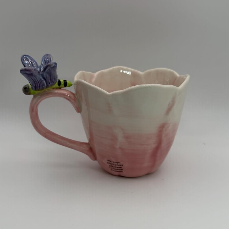 May include: A ceramic mug with a pink and white gradient design. The mug features a handle and a decorative dragonfly with purple wings and a green body. The mug has a scalloped rim.