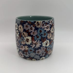 Puede incluir: Un vaso de cer&aacute;mica con un estampado floral. El vaso presenta un fondo azul oscuro con un patr&oacute;n repetido de peque&ntilde;as flores en tonos blancos, azules y marrones. El borde es de color verde azulado.