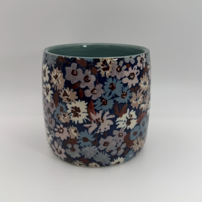 Puede incluir: Un vaso de cer&aacute;mica con un estampado floral. El vaso presenta un fondo azul oscuro con un patr&oacute;n repetido de peque&ntilde;as flores en tonos blancos, azules y marrones. El borde es de color verde azulado.