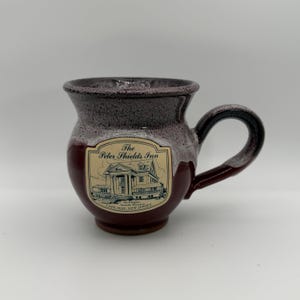 May include: A maroon and brown ceramic mug with a handle. The mug features a label with the text "The Peter Shields Inn" and an illustration of a building. The mug is a unique shape and has a glossy finish.
