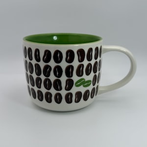 May include: White ceramic coffee mug with a green interior and a handle. The mug is decorated with a pattern of brown coffee beans. Two green coffee beans are also featured on the mug.