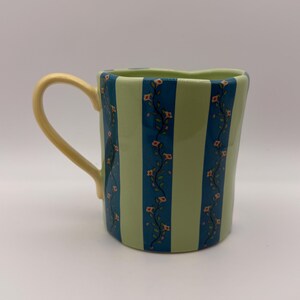 May include: A ceramic mug with a light green and blue striped pattern. The mug features a yellow handle and is decorated with floral vines. The mug is a unique shape and is suitable for hot or cold beverages.
