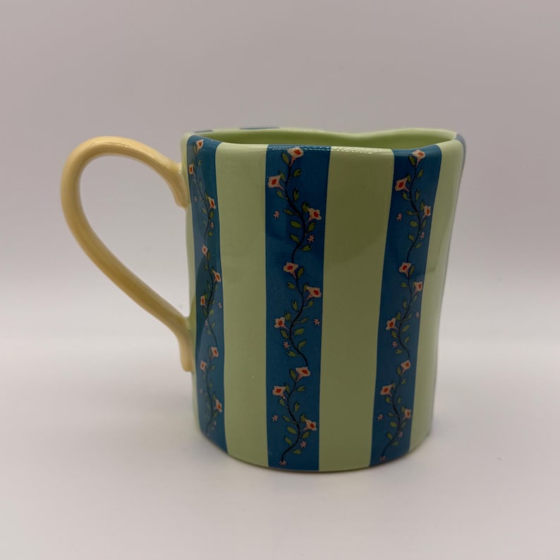 May include: A ceramic mug with a light green and blue striped pattern. The mug features a yellow handle and is decorated with floral vines. The mug is a unique shape and is suitable for hot or cold beverages.