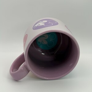 Puede incluir: Una taza de cer&aacute;mica blanca con asa e interior lavanda. El exterior presenta siluetas de cabeza de Mickey Mouse rosas y lavanda. La base interior tiene un dise&ntilde;o de cabeza de Mickey Mouse turquesa. La taza est&aacute; en &aacute;ngulo, mostrando el interior.