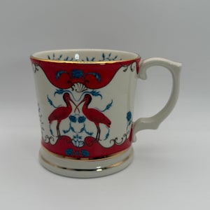 May include: A white ceramic mug with a red and blue design featuring two red birds. The mug has a gold trim and a curved handle. The design is symmetrical and ornate.