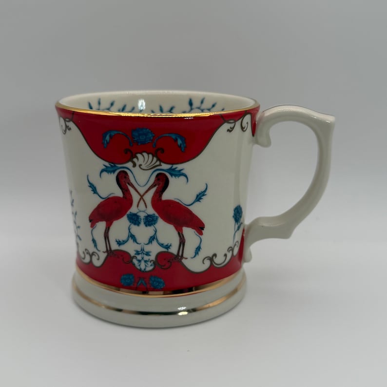 May include: A white ceramic mug with a red and blue design featuring two red birds. The mug has a gold trim and a curved handle. The design is symmetrical and ornate.