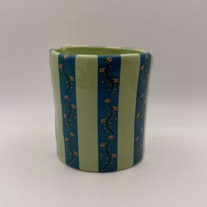 May include: A ceramic vase with a light green and teal striped pattern. Teal stripes alternate with light green stripes. Delicate floral vines with small pink flowers decorate the teal stripes. The vase has a slightly irregular shape.