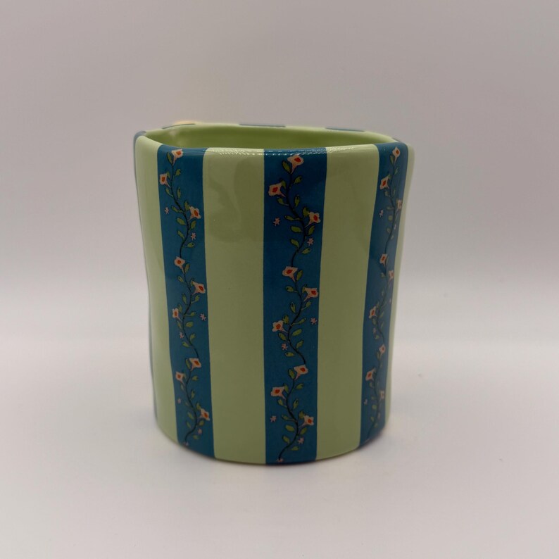 May include: A ceramic vase with a light green and teal striped pattern. Teal stripes alternate with light green stripes. Delicate floral vines with small pink flowers decorate the teal stripes. The vase has a slightly irregular shape.