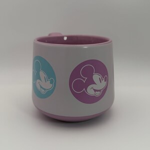 Puede incluir: Una taza de cer&aacute;mica blanca con interior y base de color lila claro. La taza presenta dos dise&ntilde;os circulares con la imagen de Mickey Mouse, uno en un c&iacute;rculo azul y otro en un c&iacute;rculo morado. El asa tambi&eacute;n es de color lila claro.
