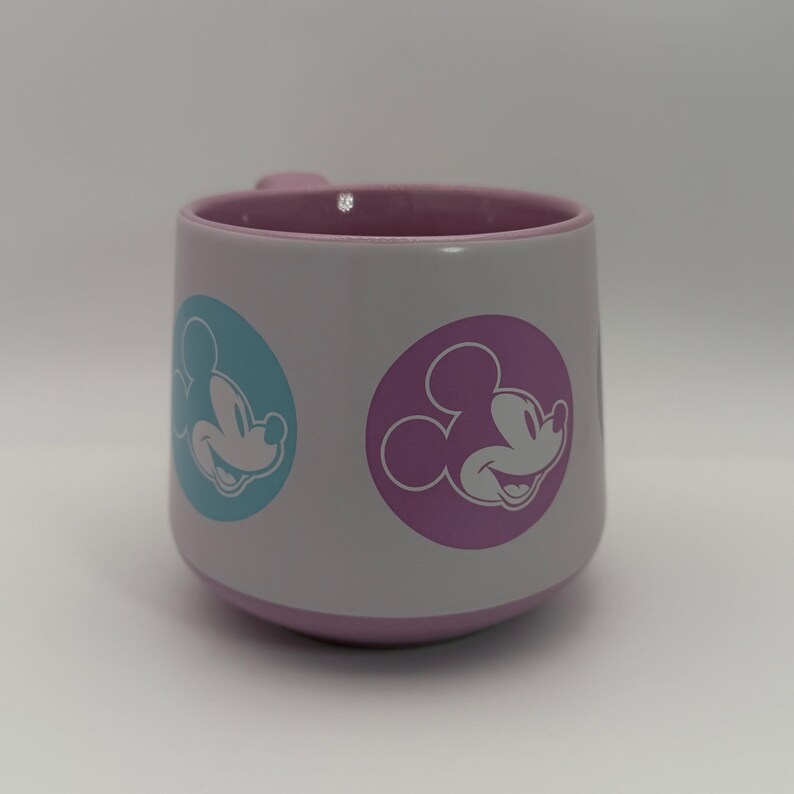Puede incluir: Una taza de cer&aacute;mica blanca con interior y base de color lila claro. La taza presenta dos dise&ntilde;os circulares con la imagen de Mickey Mouse, uno en un c&iacute;rculo azul y otro en un c&iacute;rculo morado. El asa tambi&eacute;n es de color lila claro.