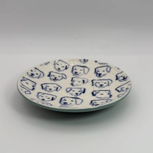 May include: A small, round ceramic plate with a white base and a blue rim. The plate is decorated with a repeating pattern of blue dog faces. The plate is likely used for serving small portions of food or as a decorative item.